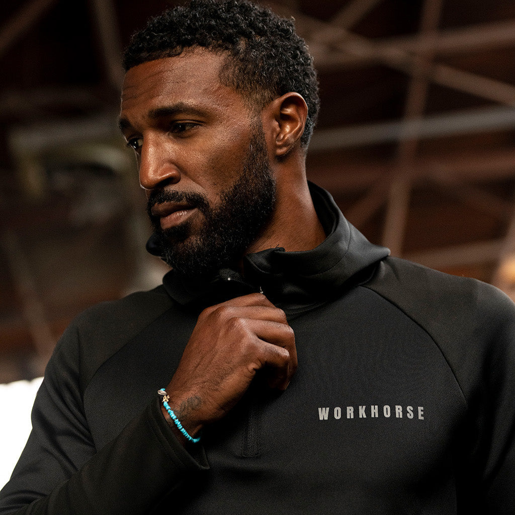 Man wearing Workhorse's High Collar Training Hoodie with logo in an industrial setting background.