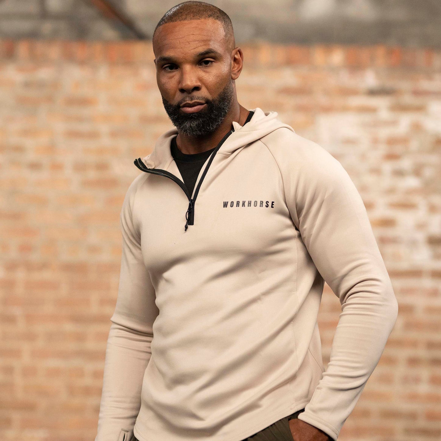 Man wearing Workhorse's High Collar Training Hoodie in sand with brand logo against a brick wall.