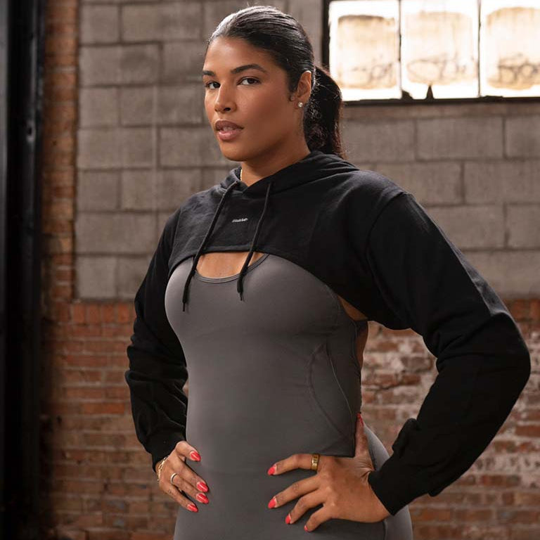 Woman wearing Mare's Crop Top Hoodie in black and gray jumpsuit standing against a brick wall.