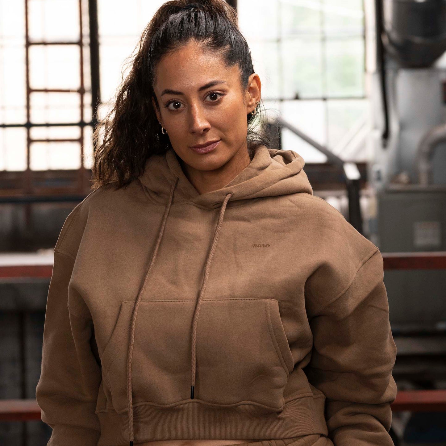 Woman wearing Mare's Hoodie in color brown and Mare Joggers in an industrial setting.