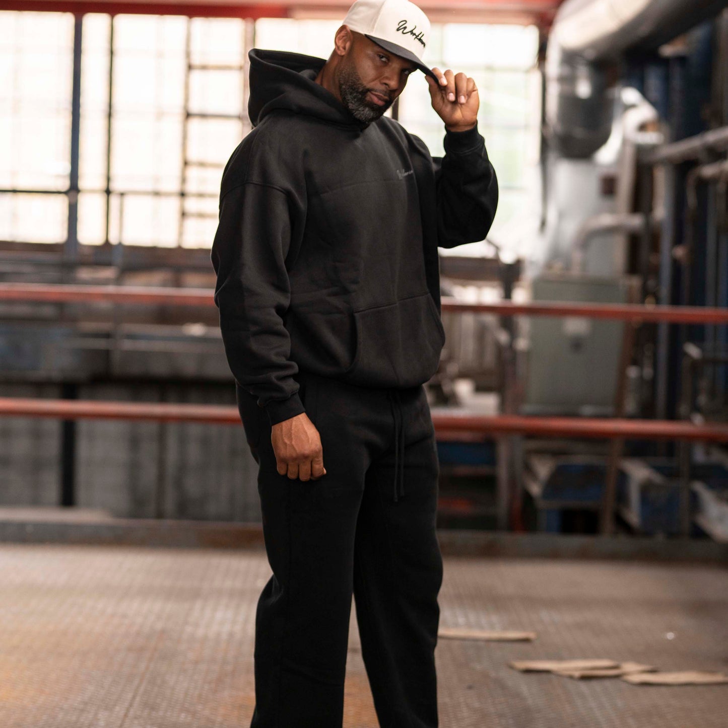 Model wearing Workhorse Oversize Hoodie and Workhorse Wide Leg Fleece Pants set in color black standing in an industrial setting.