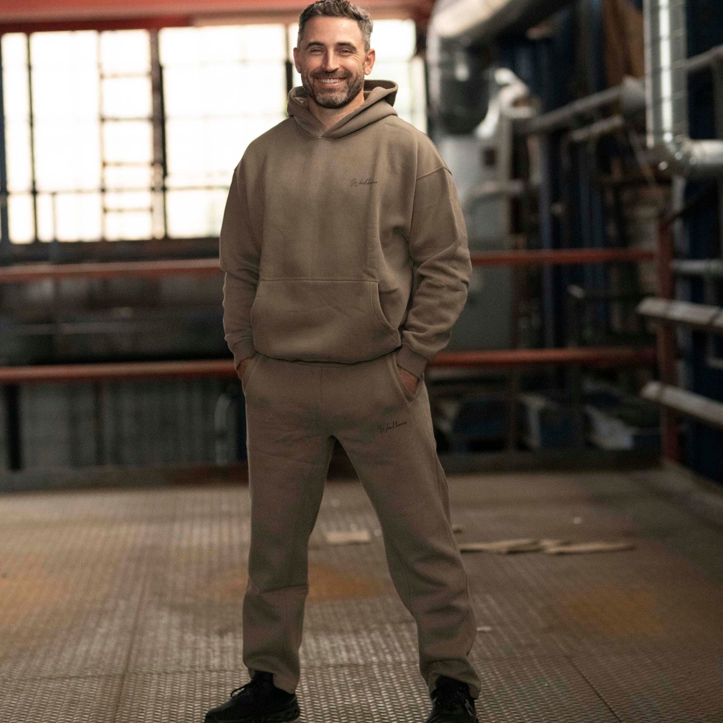 Model wearing Workhorse Oversize Hoodie and Workhorse Wide Leg Fleece Pants in color brown standing in an industrial setting.