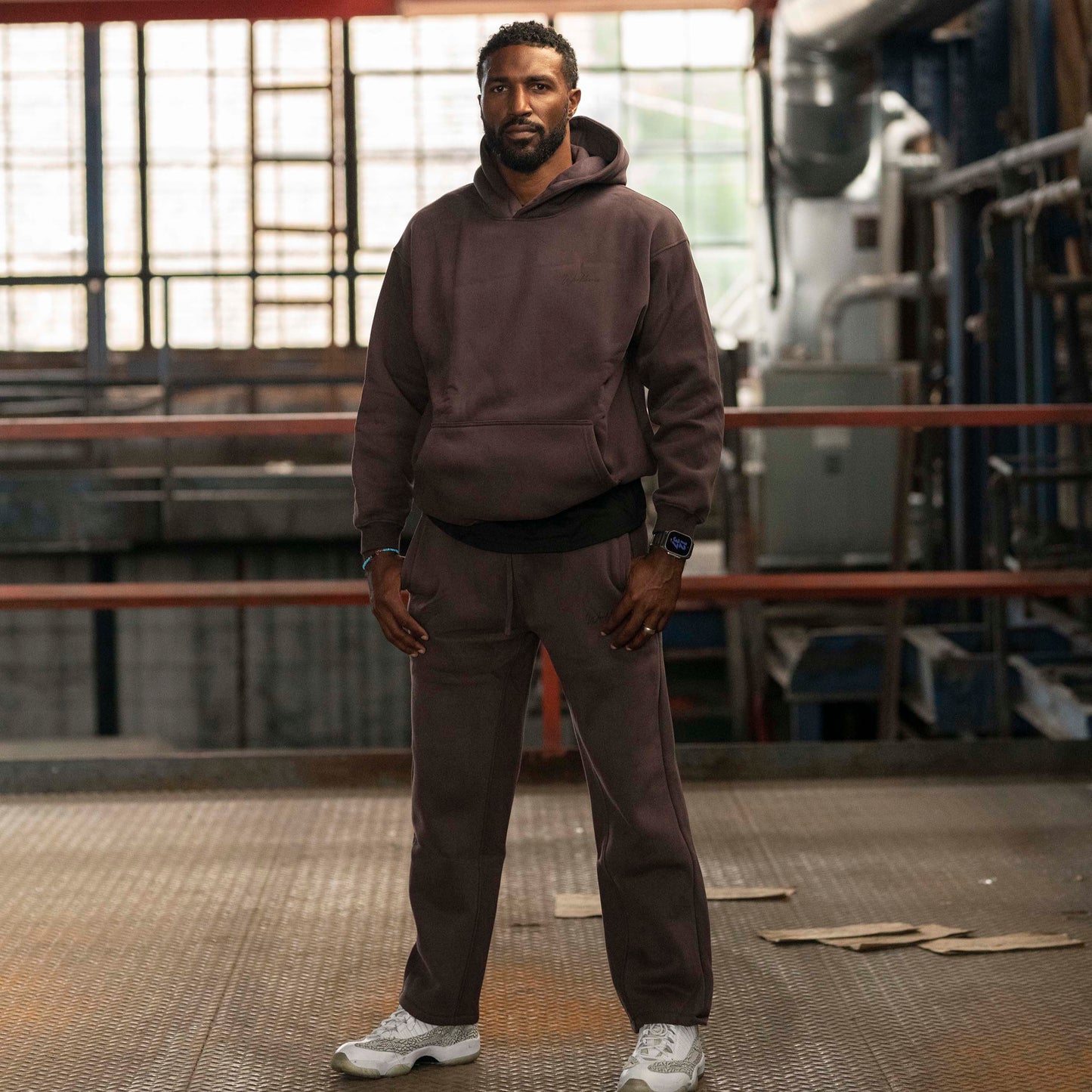 Model wearing Workhorse Oversize Hoodie and Workhorse Wide Leg Fleece Pants in color brown standing in an industrial setting.