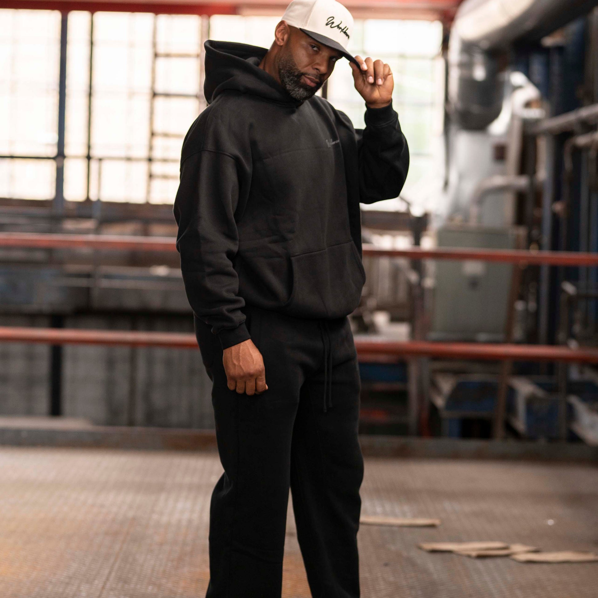 Model wearing Workhorse Oversize Hoodie and Workhorse Wide Leg Fleece Pants set in color black standing in an industrial setting.